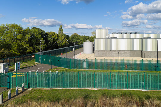 Aerial view of a modern data center facility with secure green fencing, surveillance cameras, and industrial equipment under a blue sky in the United Kingdom.