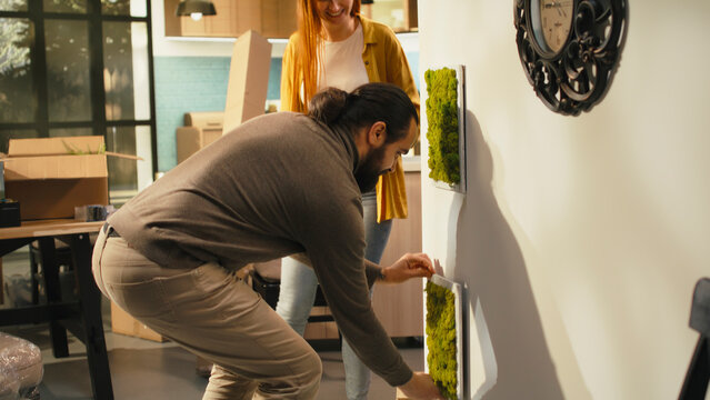 Couple arranges decorations and paintings on the wall of a new house while unpacking belongings from cardboard boxes on relocation day and creating a cozy home together for a fresh start.
