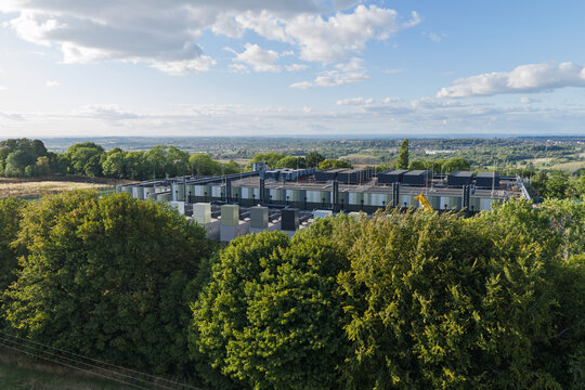 Aerial view of a large data center facility surrounded by dense green trees with a rolling landscape in the background under a blue sky in United Kingdom.