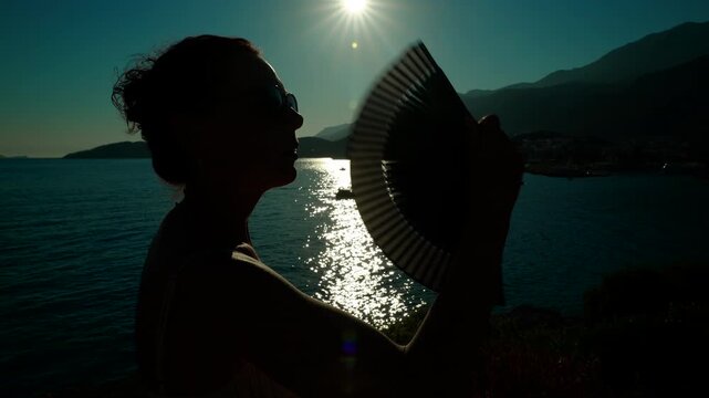 Silhouette of a woman with a fan enjoying the seaside sunset. Backlit silhouette of a woman with sunglasses and her hair in a bun fanning herself with a hand fan, enjoying the sea and the sunset