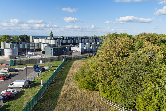 Aerial view of a modern data center facility with industrial buildings, cooling units, a parking lot, and surrounding green trees under a clear blue sky in the United Kingdom.