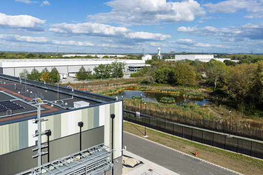 Aerial view of a modern data center facility featuring rooftop solar panels, a small pond, and surrounding greenery with a large warehouse in the background in the United Kingdom.