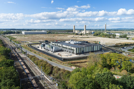 Aerial view of a modern data center facility situated next to a railway line with industrial chimneys in the background under a blue sky in the United Kingdom.