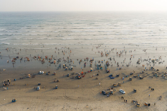 Aerial view of Clifton Beach with numerous people walking along the shoreline, scattered vehicles, and gentle waves rolling onto the sand during sunset in Karachi, Sindh, Pakistan.