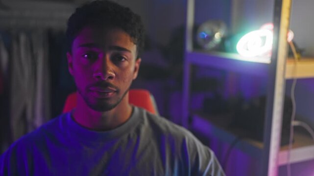 Man seated in red chair, face lit by purple neon, looking at camera in studio; focused concentration.