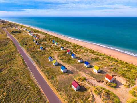 Aerial view of colorful beach huts scattered along the sandy dunes next to a coastal road overlooking the blue sea in Gouville-sur-Mer, Normandy, France.
