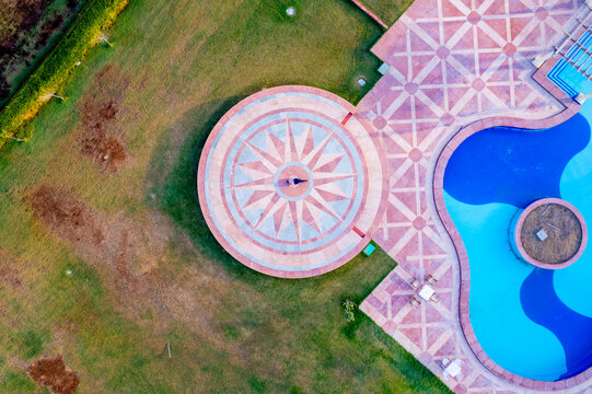 Aerial view of an elegant resort featuring a circular stone deck with traditional Rajasthani patterns, a blue swimming pool, and manicured green lawns in Jodhpur, Rajasthan, India.