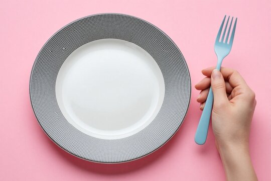 Female hand holds blue fork near empty ceramic plate on pink background. Top view close up shows woman skipping meal practicing fasting. Concept of weight loss, dieting, hunger, minimalism in