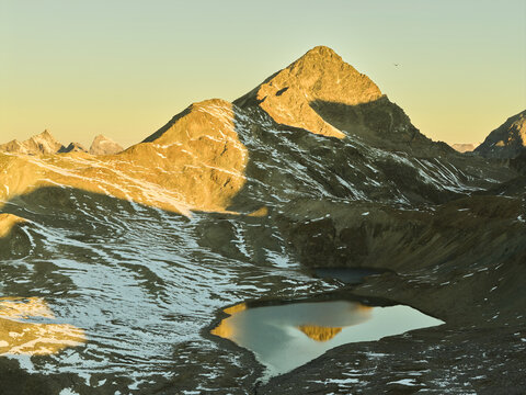 Aerial view of Engadin mountain range with snow-dusted slopes and an alpine lake reflecting the golden hour light in Grisons, Switzerland.