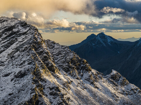 Aerial view of Valchiavenna mountains with snow-capped peaks and rocky ridges illuminated by golden hour light in Lombardia, Italia.