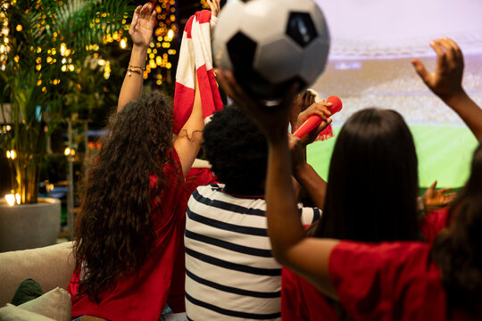 Football fans cheer at a rooftop soccer watch party, holding a soccer ball and team scarves overhead while watching the match on a projector screen at night with supporters during the competition.