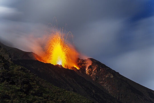 Aerial view of the Stromboli volcano erupting with glowing lava fountains and volcanic ash against a dark twilight sky, Lipari, Sicily, Italy.