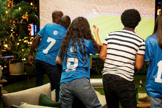 Football supporters in team jerseys watch a rooftop match projection with intense focus, following a tense counterattack on screen during a nighttime soccer watch party with friends on the terrace.