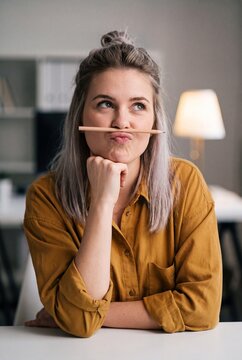 A woman with a pencil across her upper lip pouts playfully, her chin resting on her hand, in a creative and whimsical pose.