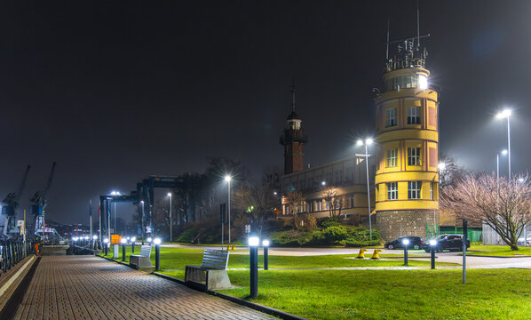View of the illuminated Gdansk Harbour Master's Office at night, featuring a paved waterfront promenade and industrial cranes in the background in Gdansk, Pomeranian Voivodeship, Poland.