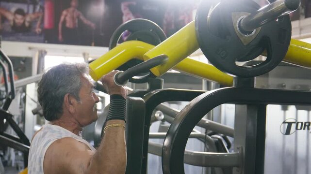 Indian Asian fit muscular elderly man using a lat pulldown machine to perform a resistance strength training exercise targeting back and shoulders in modern gym. Fitness, workout, bodybuilding concept