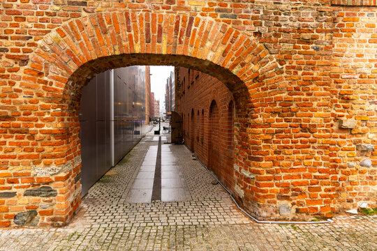 View of a historic red brick archway leading into a narrow cobblestone alleyway with modern architecture in the background in Gdansk, Pomeranian Voivodeship, Poland.