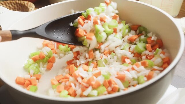 Cook uses a black silicone spatula to mix chopped onions, carrots, and celery mirepoix base in a hot skillet. Healthy vegetable preparation process for delicious homemade one-pot rice recipe.