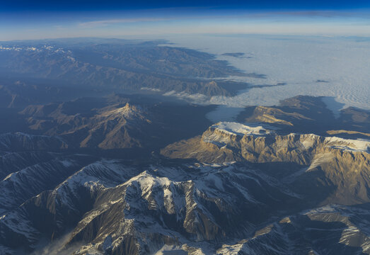 Aerial view of the snow-capped Caucasus Mountains featuring rugged peaks, deep valleys, and a layer of clouds stretching toward the horizon under a clear blue sky, Azerbaijan.