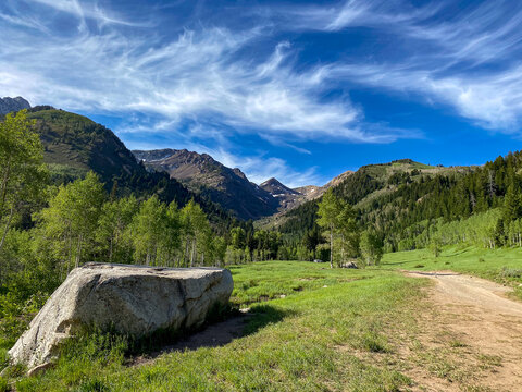 View of Uinta National Forest with a large boulder, dirt road, green meadow, and aspen trees set against mountains under a blue sky in Utah County, Utah, United States.