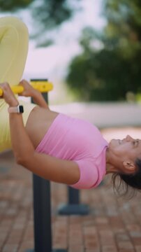 Upside-down female athlete gripping yellow parallel bars in outdoor calisthenics park, smartwatch on wrist, pink sports top and terracotta pavers with brick posts and leafy trees