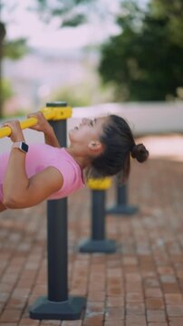 Young woman performing inverted row on bright yellow parallel bars outdoors, pulling torso upward in low diagonal view highlighting grip and arm strength