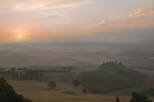 View of rolling hills of Tuscany at sunset with cypress trees and farmhouses under a soft golden sky, Tuscany, Italy.