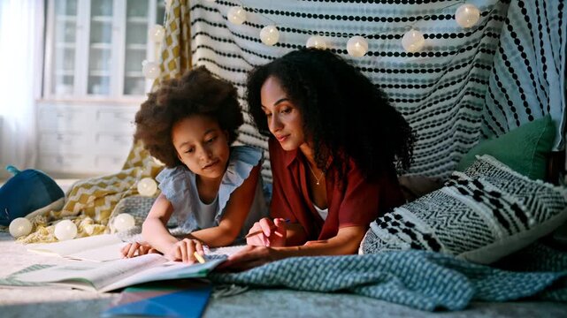 Family homeschooling blanket fort bedroom. Mom teaching daughter write notebook.