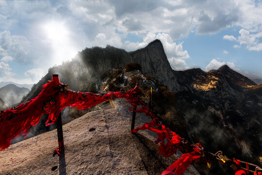 View of the steep granite peaks of Mount Hua adorned with traditional red prayer ribbons tied to iron chains under a bright sun in Xi'an, Shaanxi, China.