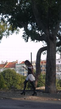 Woman walking along shaded park path beside tall trees in urban neighborhood, black heeled boots and dark top with white wrap skirt, city rooftops and pale sky in background