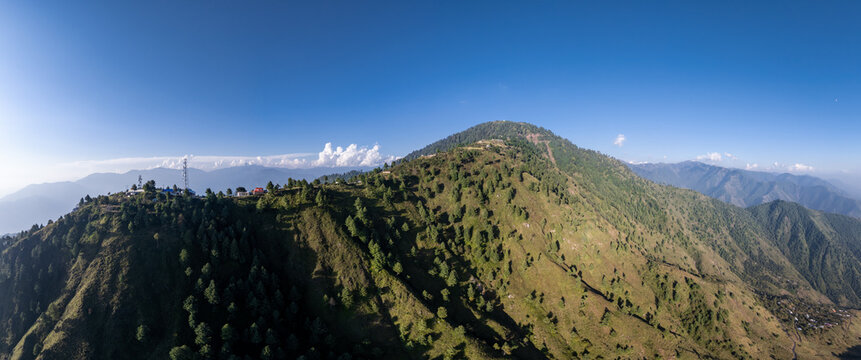 Aerial view of the Pir Chanasi mountain ridge featuring lush green slopes, scattered pine trees, and communication towers under a clear blue sky in Pir Chanasi, Azad Kashmir, Pakistan.