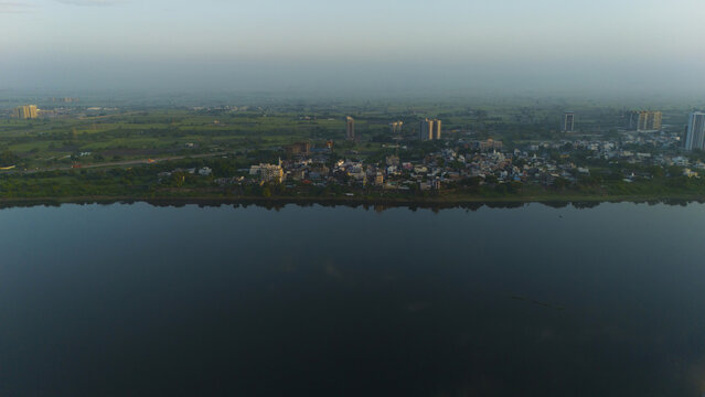 Aerial view of the Tapi River flowing past the urban landscape with scattered buildings, lush green fields, and a hazy sky at dawn in Surat, Gujarat, India.