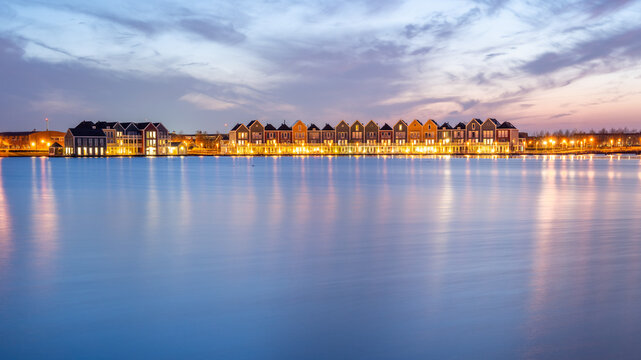 View of colorful modern houses along the calm water of a lake at dusk with warm lights reflecting on the surface in the Netherlands.