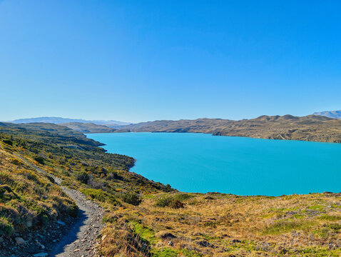 View of Torres del Paine National Park with a hiking trail leading towards the turquoise waters of the lake under a clear blue sky in Magallanes, Chile.