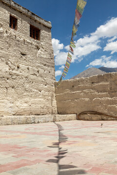 View of Charasa Gonpa with colorful prayer flags hanging against a bright blue sky and textured stone walls in a paved courtyard in Charasa, Ladakh, India.