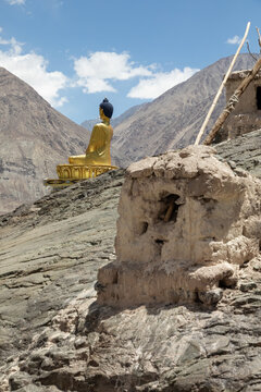 View of the golden Buddha statue at Charasa Gonpa, set against a backdrop of rugged mountains and ancient stone ruins in the arid landscape of Charasa, Ladakh, India.