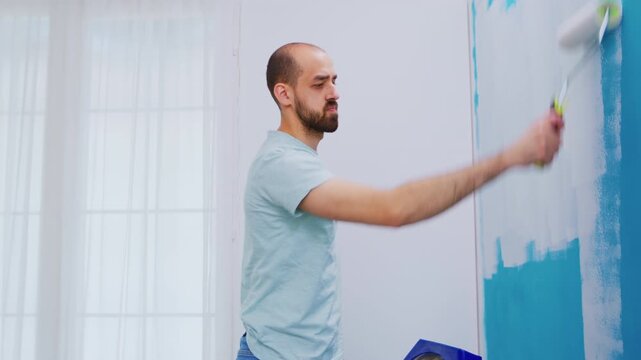 From the side, a man covers blue wall paint with white using a roller, focusing on interior repainting, color change and practical home improvement.