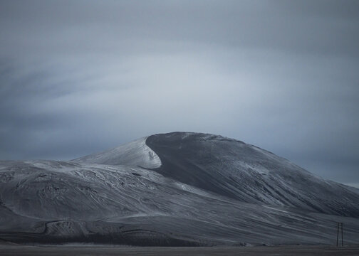 View of a snow-covered volcanic mountain with dark basaltic slopes under a dramatic overcast sky in Vik, Myrdalshreppur, Iceland.