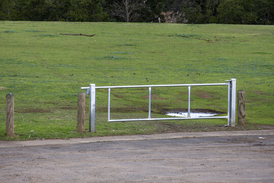 A galvanized metal swing boom gate at edge of a paved surface leading into a green parkland, flanked by wooden bollards in Australia.&nbsp;Blocking unauthorized&nbsp;vehicles from entering lawns, damaging turf