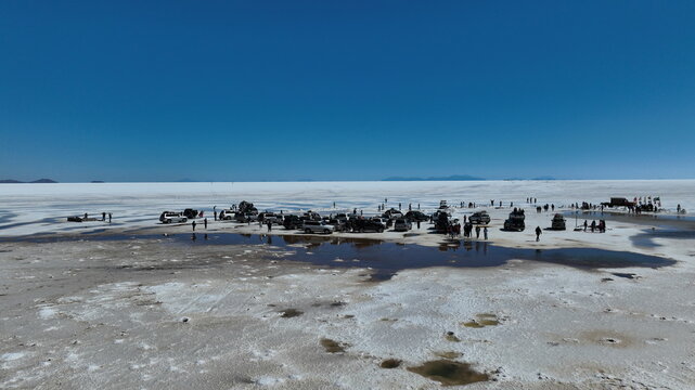 Aerial view of Uyuni Salt Flat with parked 4x4 vehicles, tourists exploring the vast white salt crust, and shallow pools of water under a clear blue sky, Potosi Department, Bolivia.