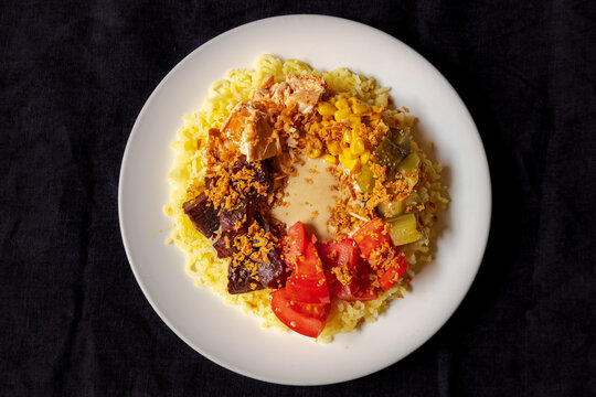 View of a plate of yellow rice topped with chicken, corn, pickles, tomatoes, and crispy fried onions arranged in a circular pattern on a white plate in Latvia.