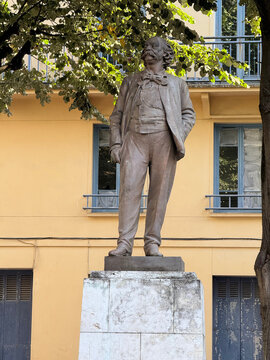 Statue of Gustave Flaubert on a stone pedestal beneath leafy trees in front of a yellow building - Rouen, France - September 6, 2026