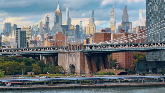 New York, USA, 9 Jule 2026 : Manhattan Bridge anchorage with the Midtown skyline behind it. A masonry arch and steel deck rise above a waterfront road, with clustered towers beneath layered clouds.