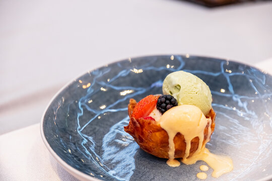 View of a gourmet dessert in a crispy pastry bowl with scoops of pistachio and vanilla ice cream, garnished with fresh strawberry and blackberry on a blue patterned plate in Latvia.