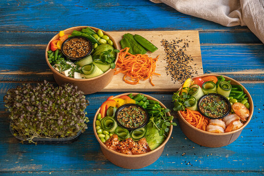 View of three colorful poke bowls with salmon, shrimp, tofu, and fresh vegetables served on a rustic blue wooden surface, Latvia.