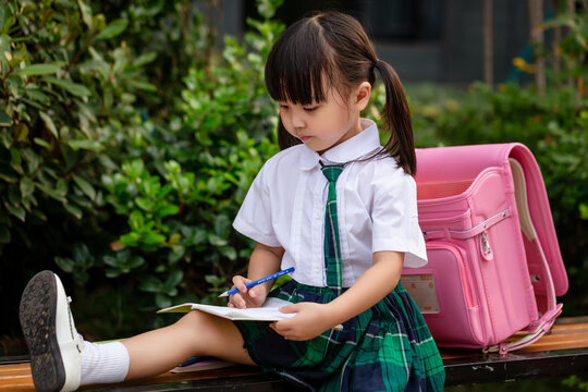 Diligent Asian girl student sitting on a bench with her backpack, writing and learning outdoors.