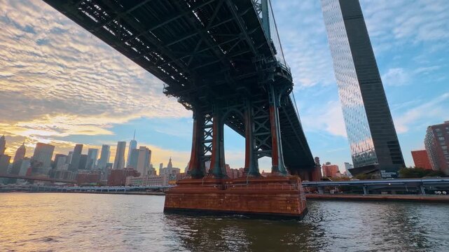 New York, USA, 9 Jule 2026 : Manhattan Bridge toward the waterfront at sunset. Dark steel beams span overhead above a masonry pier, with rippled water and a glass tower beside the shoreline.