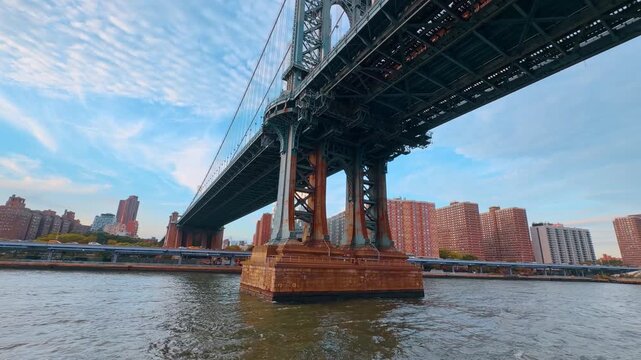 Manhattan Bridge underside and tower above the East River. A steel framework rests on a masonry pier, with rows of brick apartment buildings along the opposite bank.