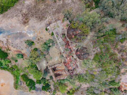 Aerial view of the abandoned mining site with rusted industrial structures, circular foundations, and overgrown vegetation surrounded by rocky terrain in Rio Marina, Toscana, Italy.