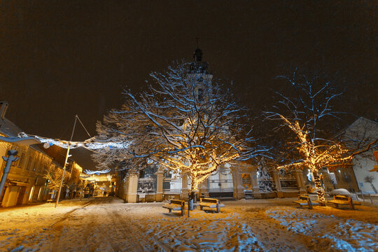 View of the Saint Demetrius Church illuminated at night during a winter snowfall with decorative fairy lights on trees in the town square of Sremska Mitrovica, Vojvodina, Serbia.